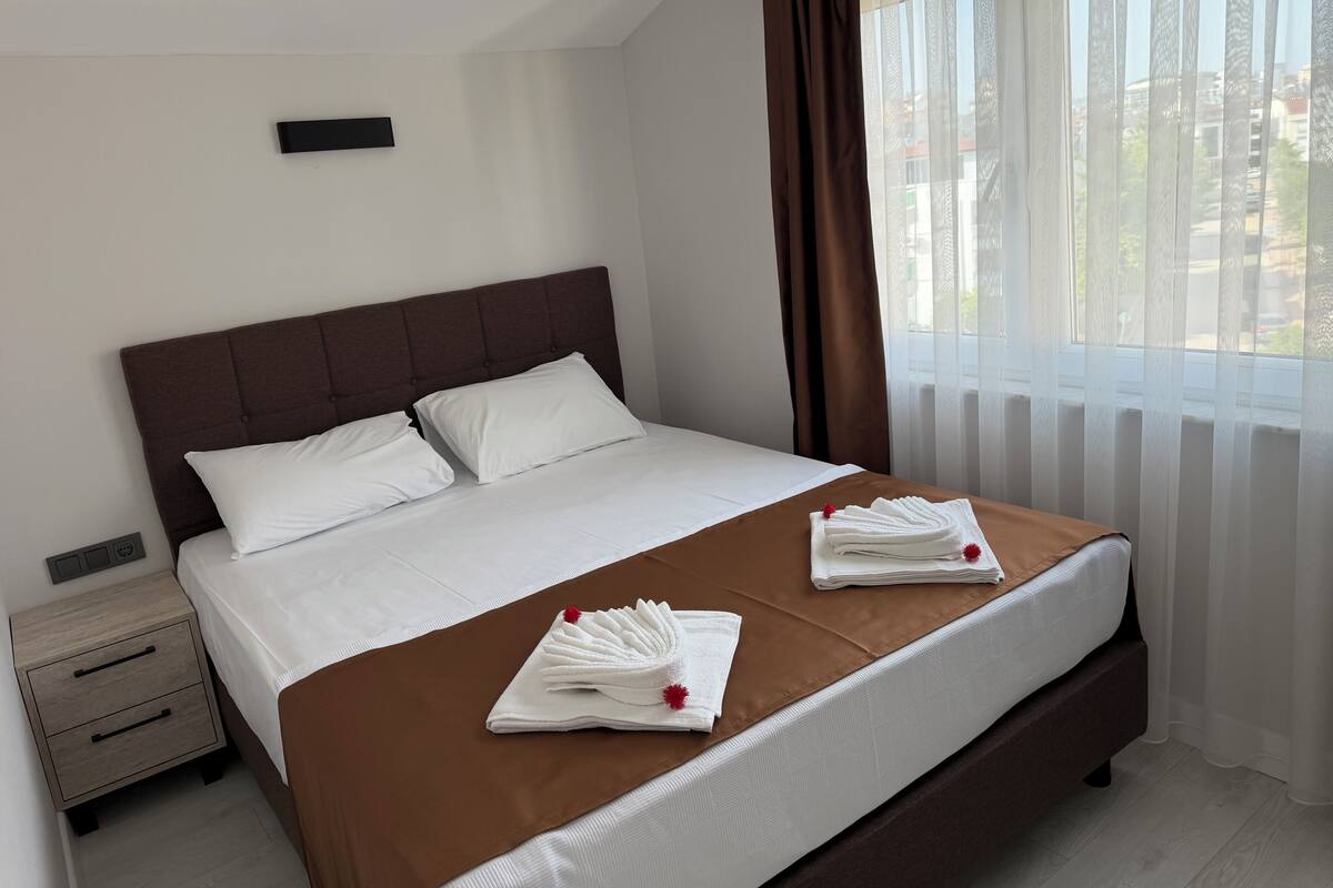 Ein ordentlich bezogenes Bett mit weißen Laken und einer braunen Tagesdecke, ein kleiner Nachttisch mit Schubladen und ein Fenster mit durchsichtigen Vorhängen.