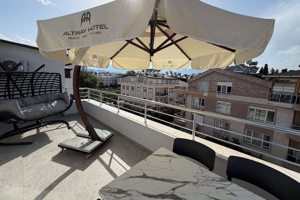 Eine Dachterrasse mit Sitzgelegenheit, inklusive Tisch und Stühlen, unter einem großen Sonnenschirm mit dem Logo des Altinay Hotels.