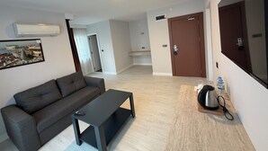 Una sala de estar moderna con sofá, una mesita de centro, una zona de cocina con hervidor y una puerta que da a otra habitación.