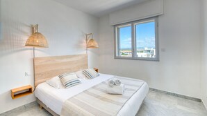 Un dormitorio moderno con cabecera de madera, cama grande con ropa de cama a rayas, dos lámparas fijadas a la pared y una ventana con vista a la ciudad.