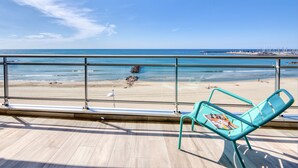 Un balcón con una silla turquesa y una revista, con vista a una playa y un mar tranquilo bajo un cielo despejado.