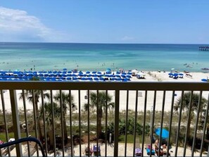A beach view from a balcony with blue umbrellas and palm trees.