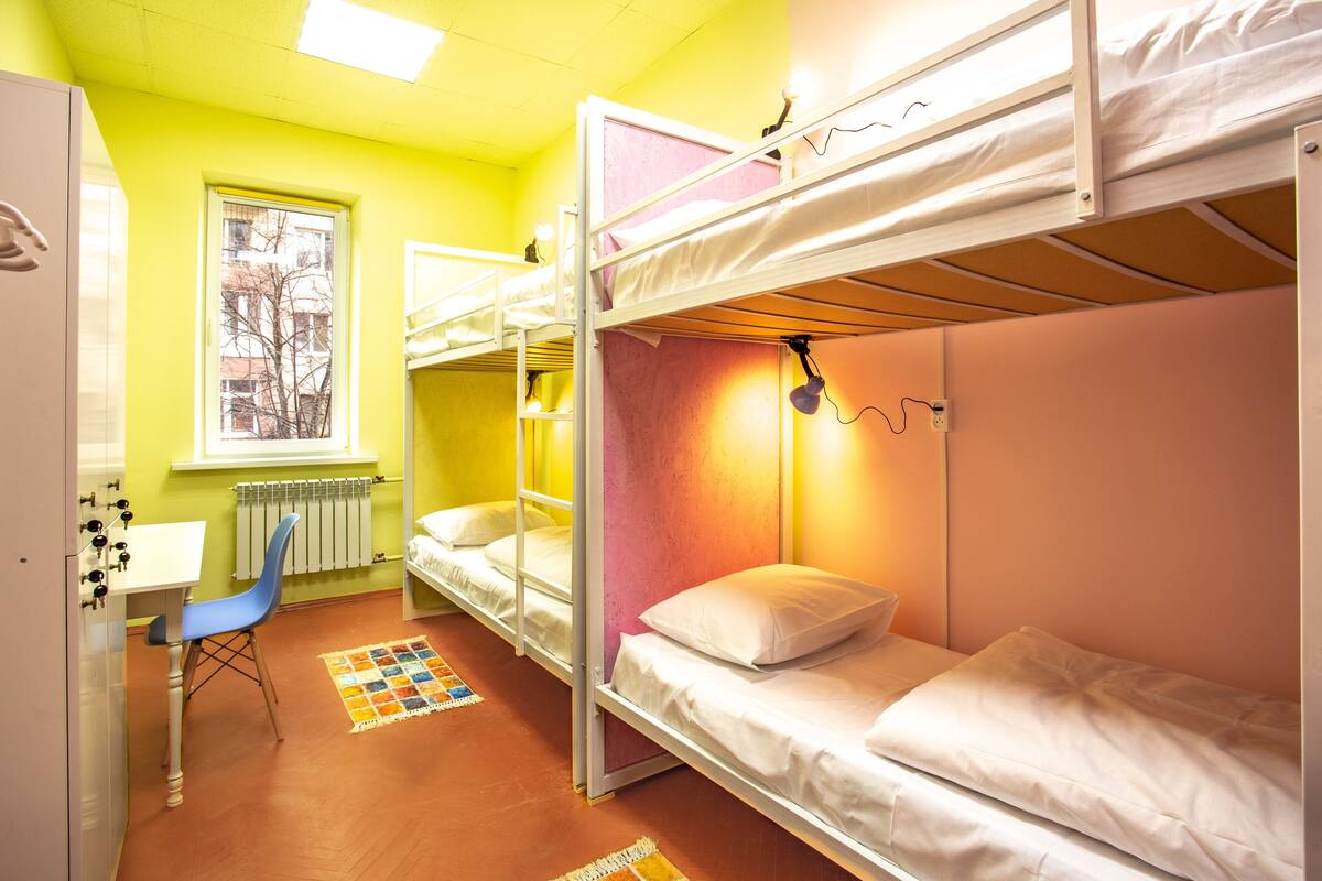 A dormitory room with bunk beds, a desk, a chair, and a window with a view of buildings.