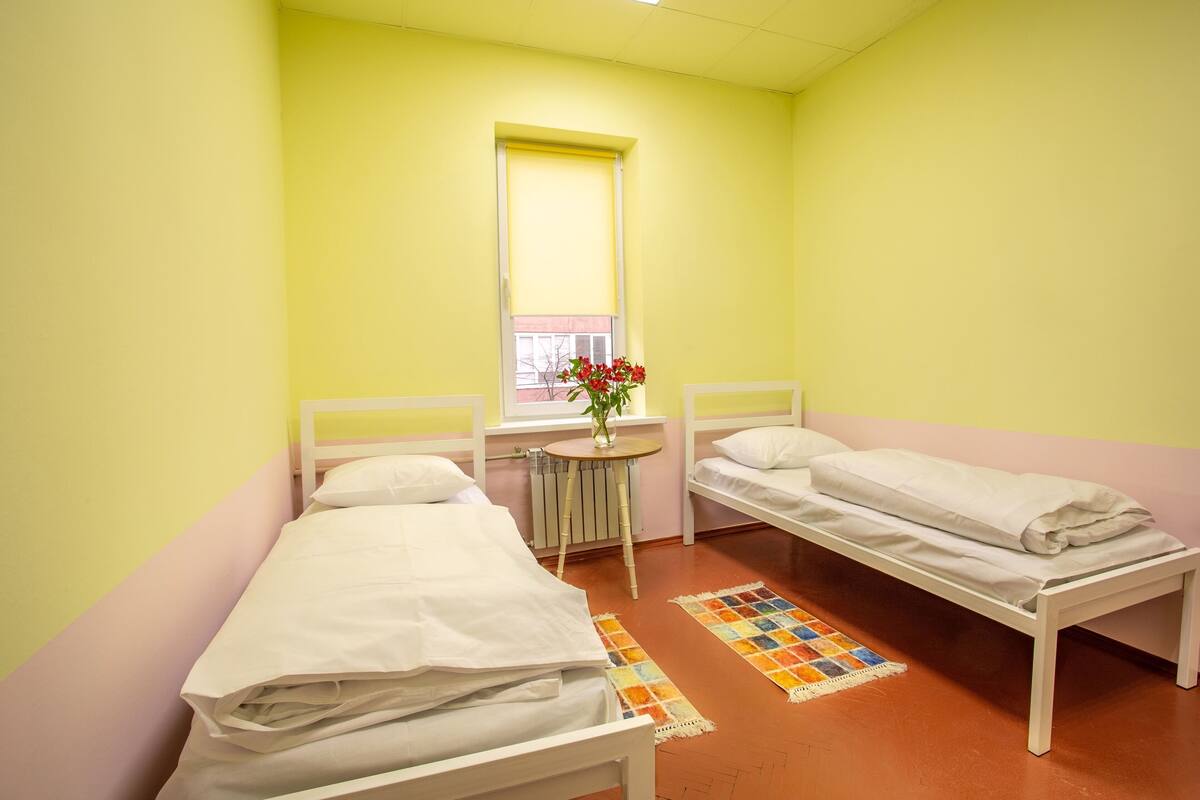 A room with two single beds, a small round table with a vase of flowers, and a window with blinds.