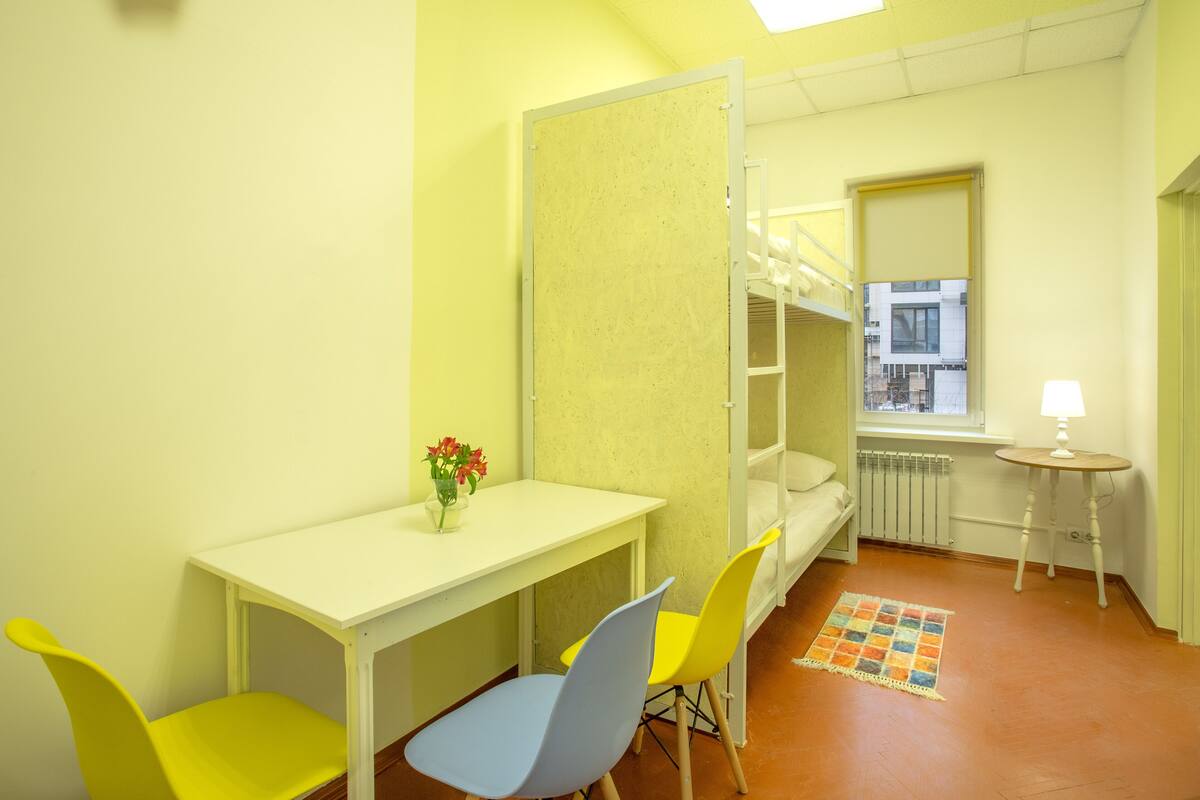 A room with a bunk bed, a desk, two chairs, a small table with a lamp, and a window with a view of buildings.