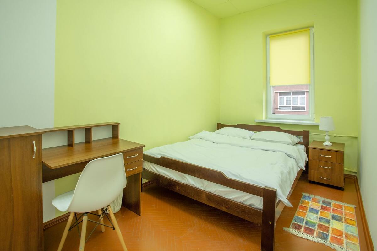 A bedroom with a bed, a desk, a chair, and a small bedside table with a lamp.