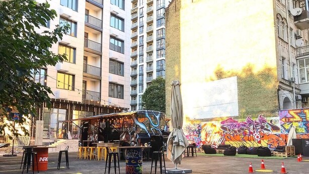 Un entorno urbano al aire libre con un puesto de comida callejera, un área de asientos y un gran mural en un edificio.