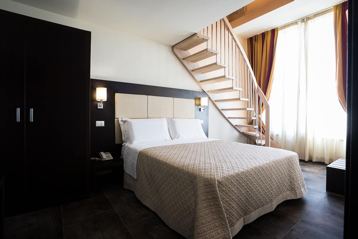 Hotelkamer met een groot bed, een donkere kast, een nachtkastje met telefoon, een wandlamp en een houten trap.