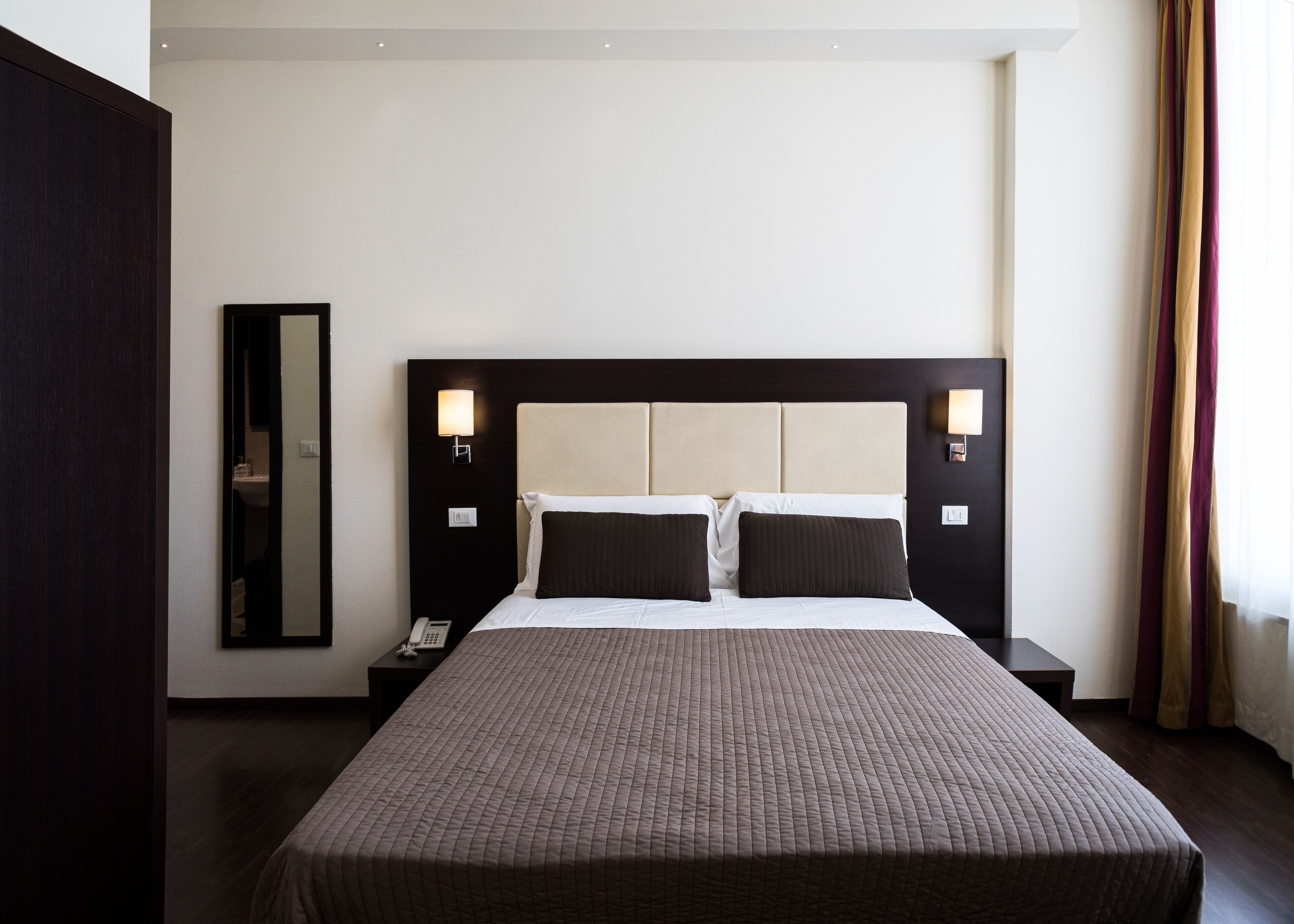 A hotel room with a large bed, two bedside tables, a nightstand, a mirror, and a window with curtains.