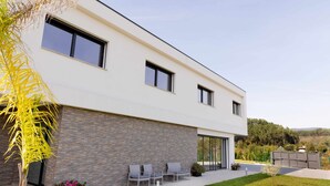 Une maison moderne à deux étages, dotée d’une façade en pierre, de grandes fenêtres et d’un patio aménagé avec du mobilier d’extérieur.
