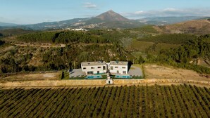 Un bâtiment moderne à deux étages, doté d’une piscine et d’un court de tennis, entouré de vignobles et surplombant un paysage montagneux.