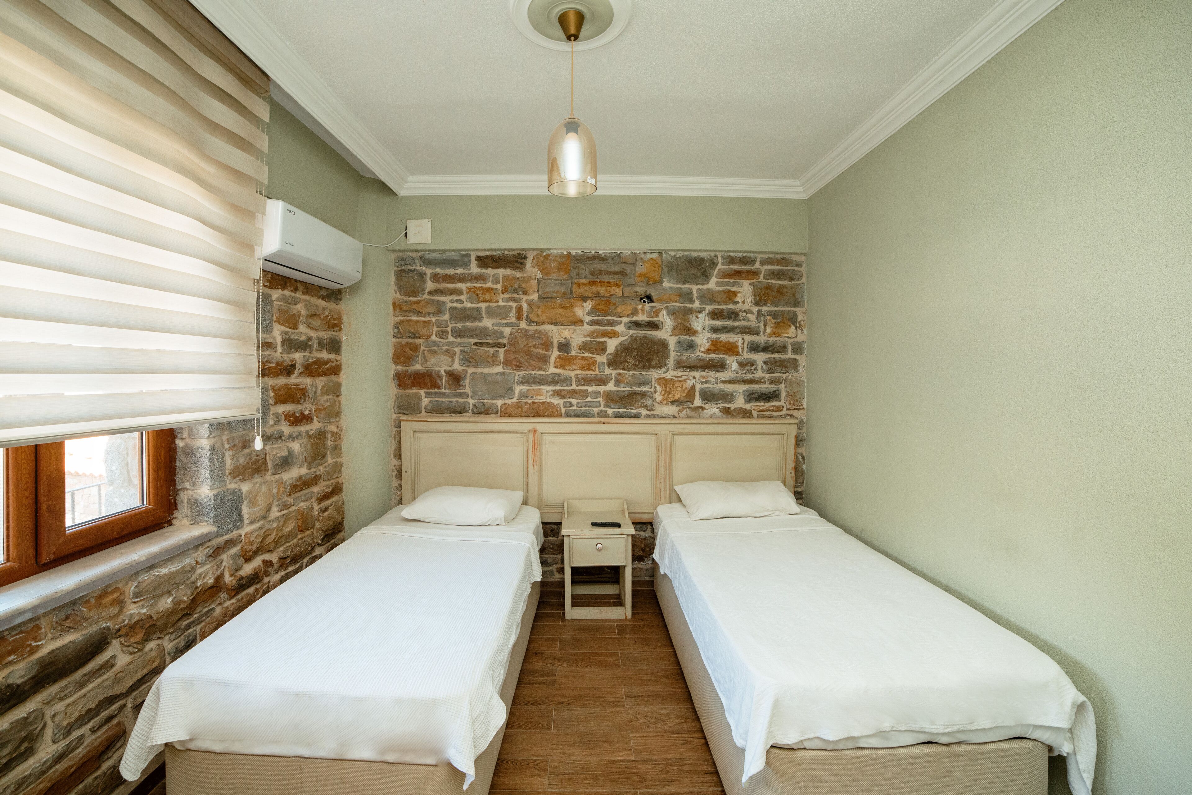 A room with two single beds, a stone wall, a window with blinds, and a hanging light fixture.