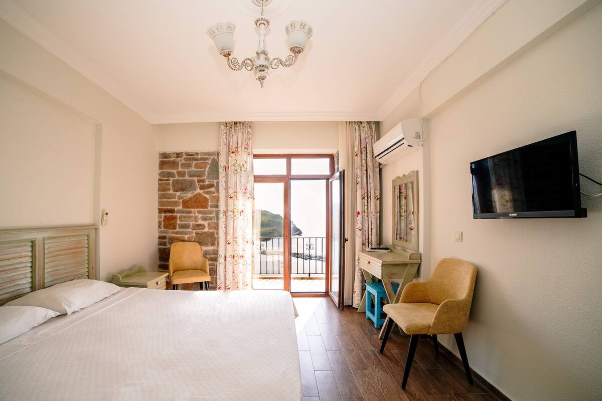 A bedroom with a bed, two chairs, a desk, a TV, and a balcony with a stone wall.