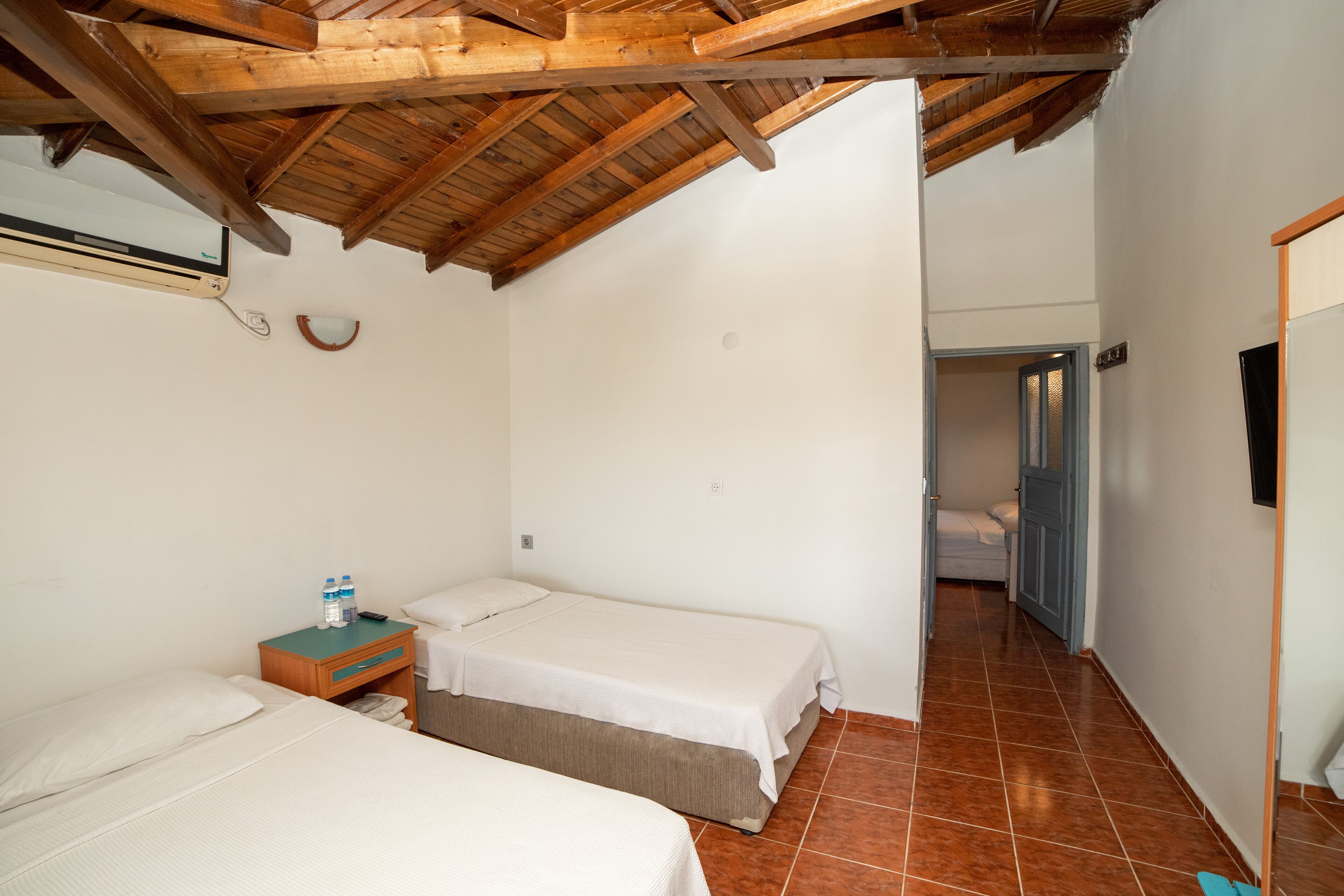 Une chambre avec deux lits simples, un plafond en bois, un sol carrelé et une porte donnant sur une autre pièce.