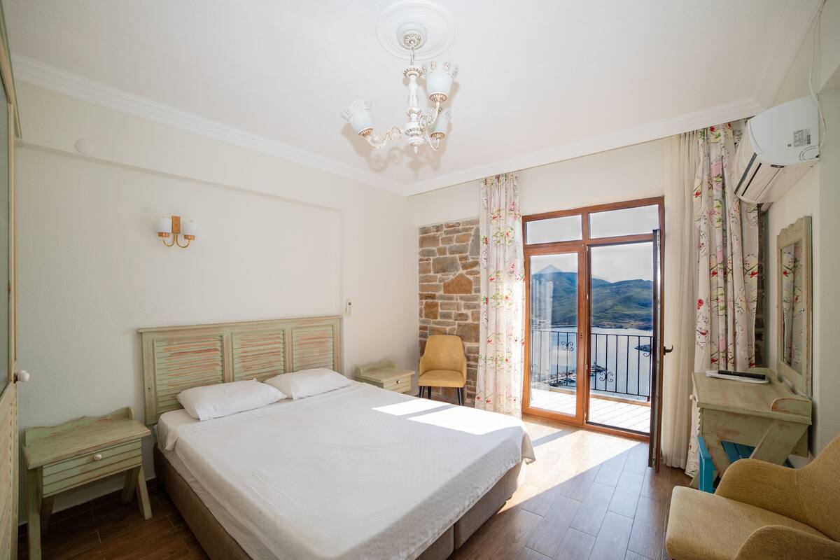 A bedroom with a large bed, a chair, a nightstand, a dresser, a balcony with a view of the mountains, and an air conditioning unit.