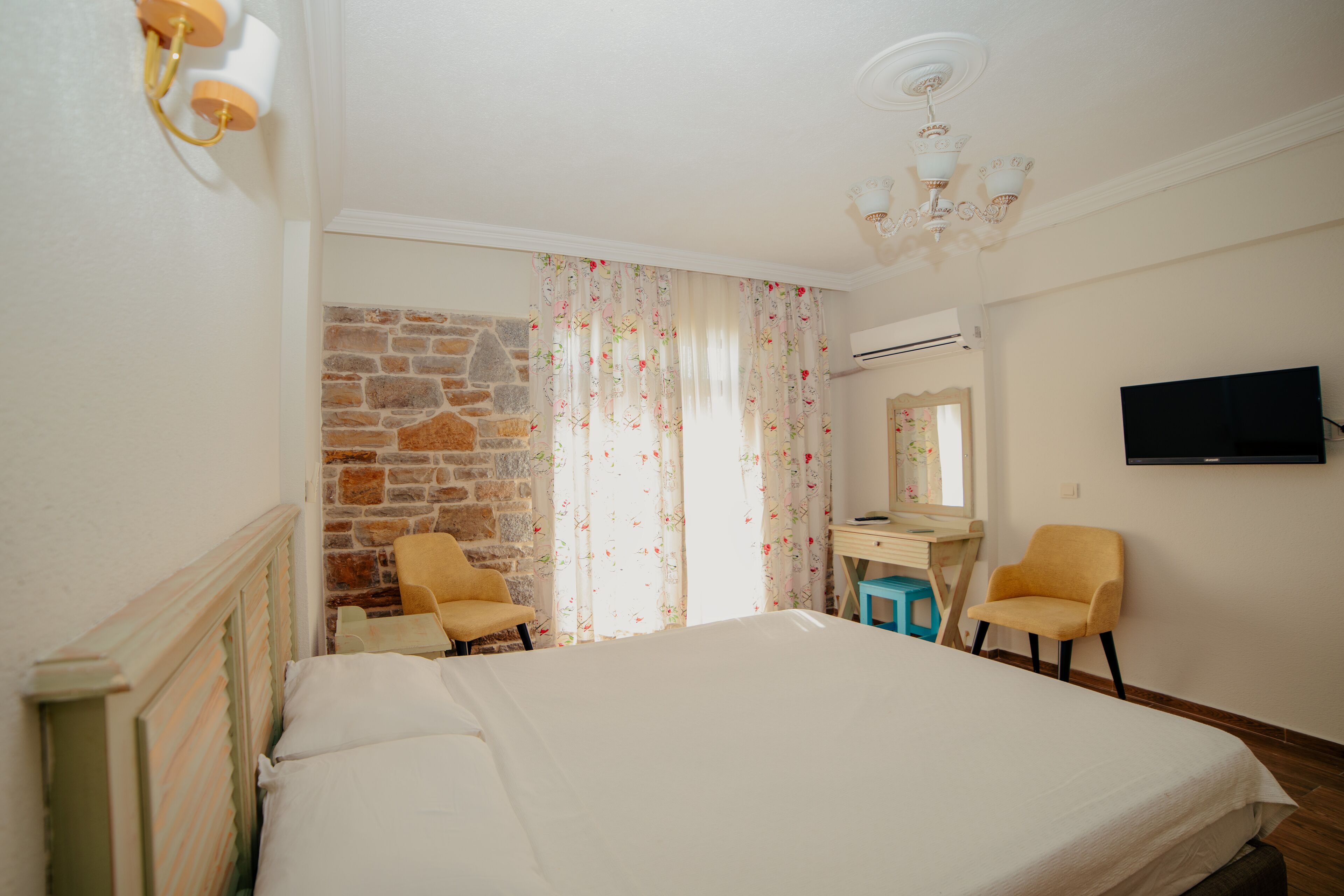 Un dormitorio con cama, dos sillas, escritorio, televisión y pared de piedra.