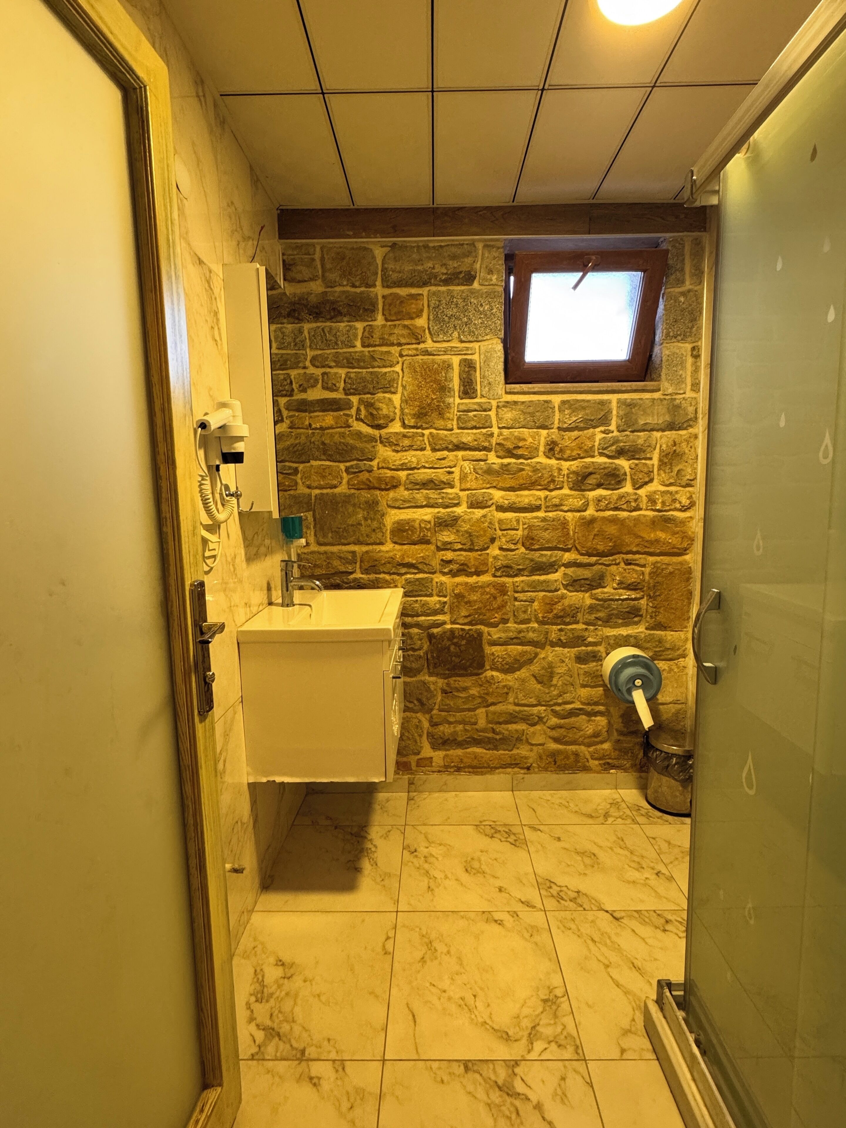 Baño con pared de piedra, una ventana pequeña, lavamanos y zona de ducha.