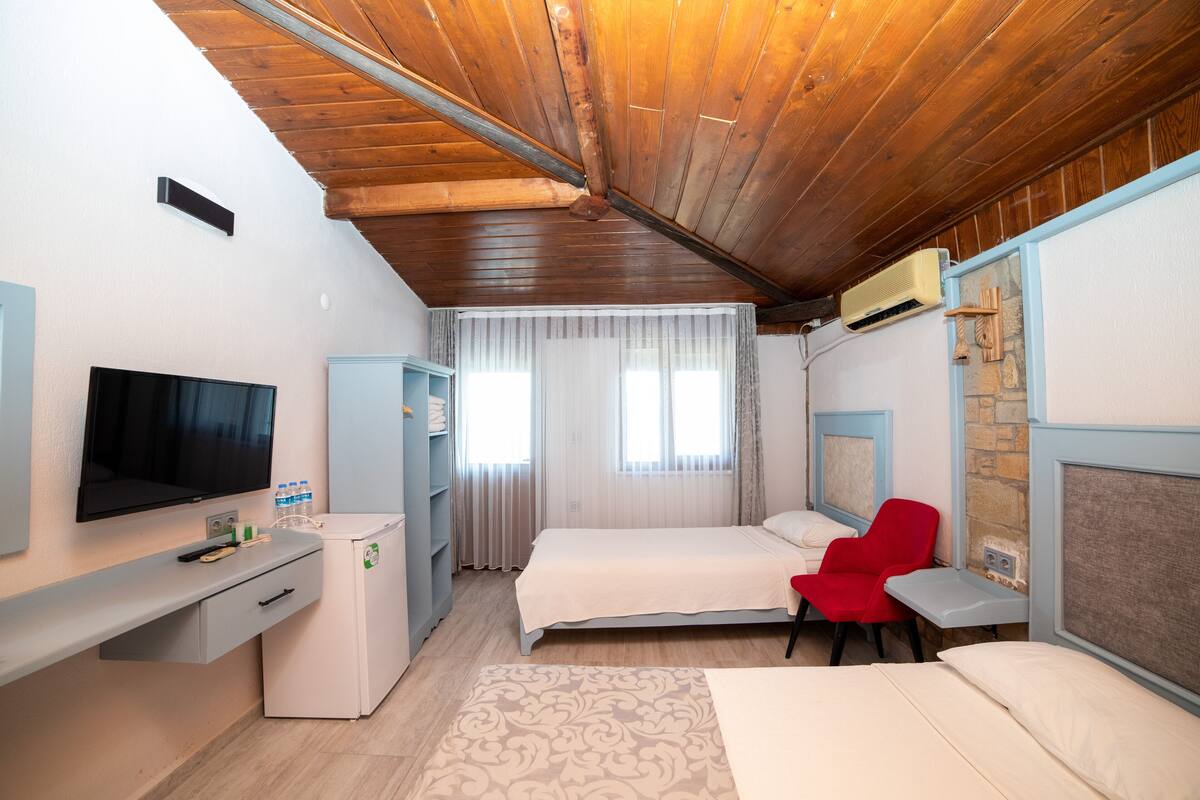 A hotel room with a wooden ceiling, a bed, a desk with a TV, a red chair, and a window with curtains.