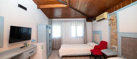 Habitación de hotel con techo de madera, una cama, un escritorio con televisor, una silla roja y una ventana con cortinas.