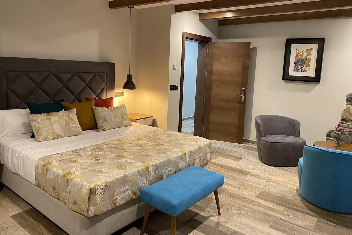 A modern bedroom with a large bed, a bench, a chair, and a wooden door.