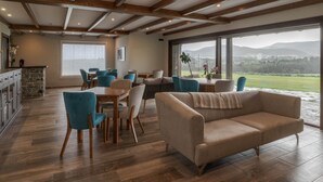 Amplia sala de estar con un ventanal que ofrece vistas a un paisaje verde y montañas. Hay un sofá color crema y varias sillas dispuestas alrededor de una mesa de comedor de madera.