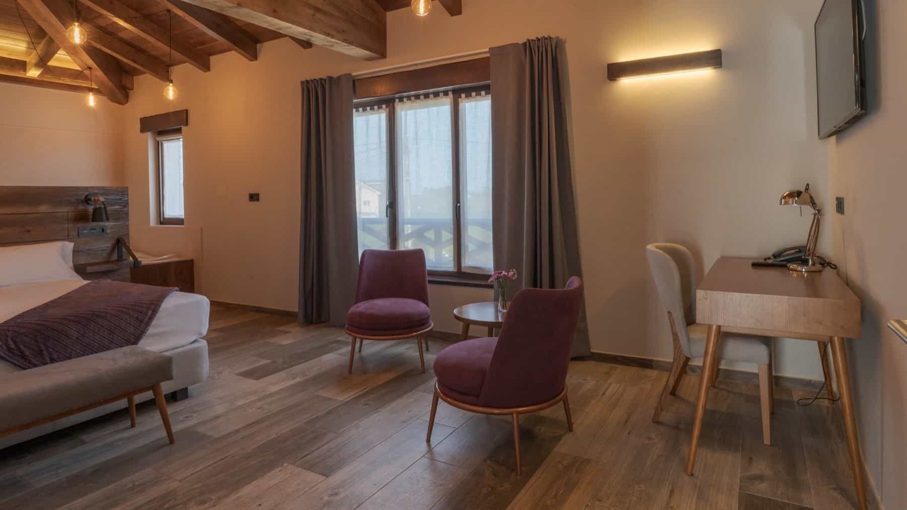 A modern hotel room with a wooden ceiling, a bed, a desk with a chair, two armchairs, and a window with curtains.