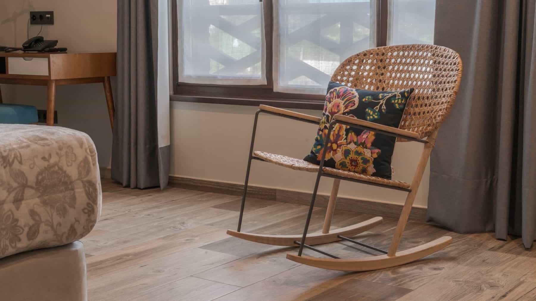 Een kamer met een houten schommelstoel, een bed met bloemenbedrukking, een bureau met een telefoon en een raam met gordijnen.