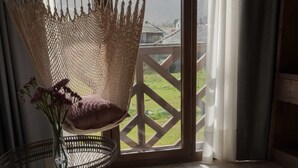 Una silla de mimbre con un cojín, un florero con flores y vistas a una terraza de madera y follaje a través de una ventana.