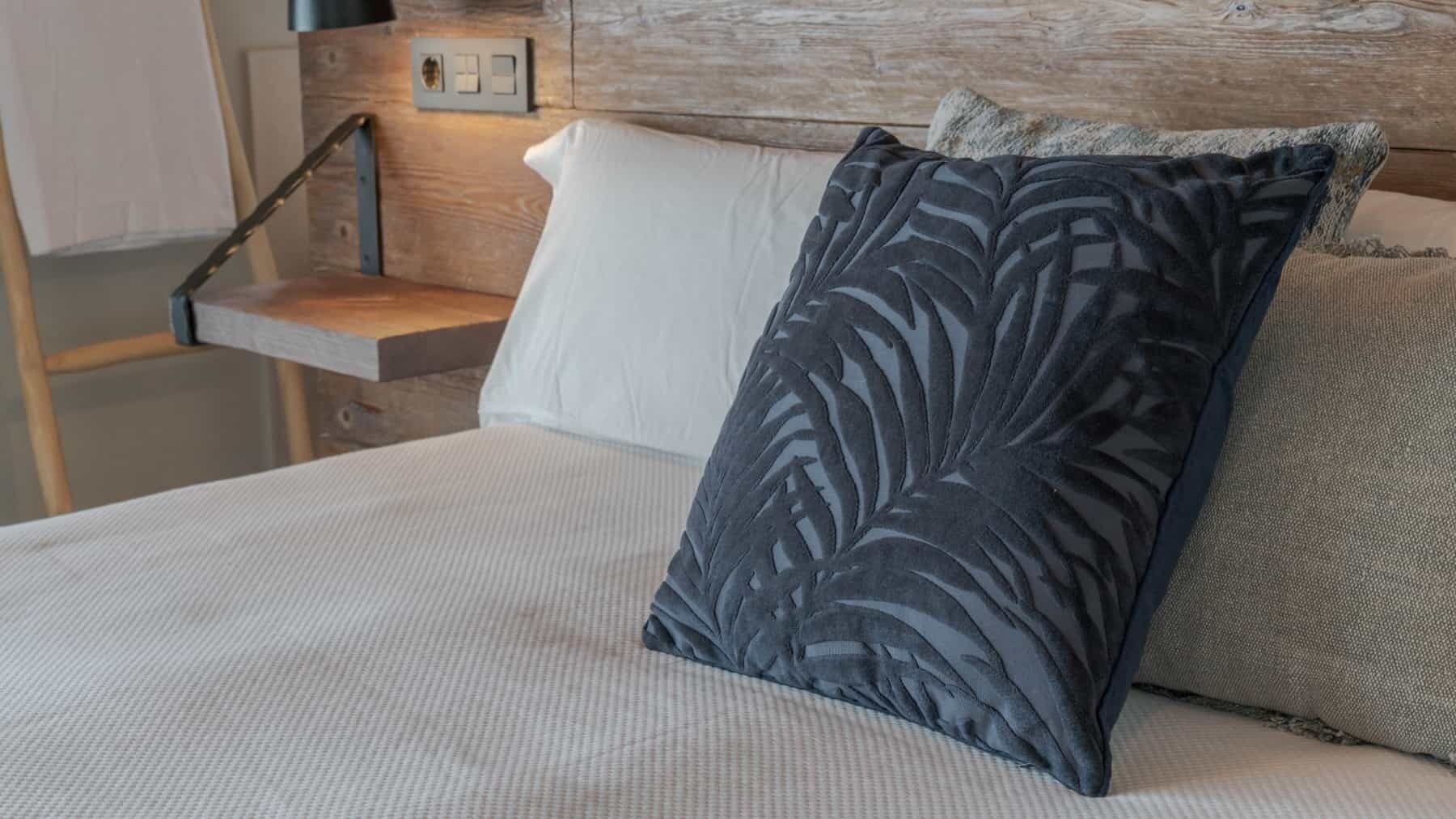 A bed with a white mattress, a dark blue pillow with a leaf pattern, and a grey textured pillow.