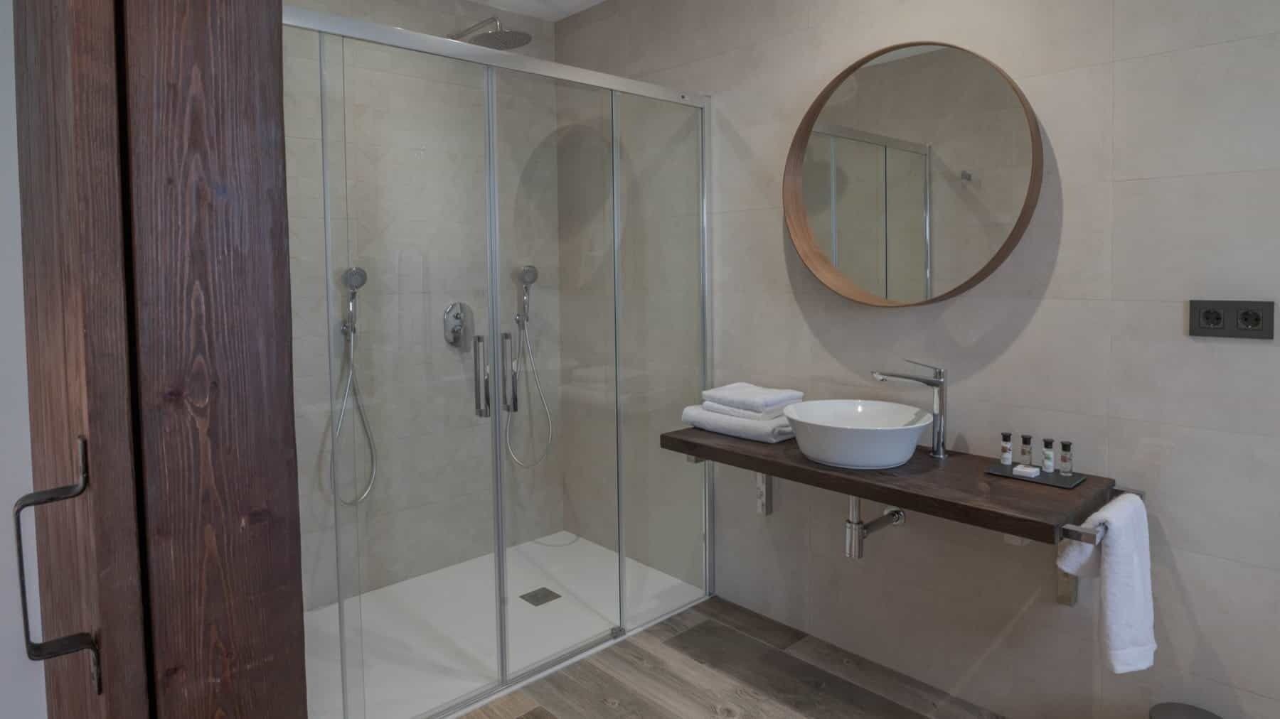 A modern bathroom with a glass-enclosed shower, a round mirror, and a white sink with a wooden countertop.