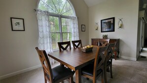 A dining area with a wooden table and chairs, a large arched window with curtains, and framed botanical prints on the walls.