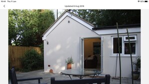 Una casita blanca con puerta de cristal que da al interior, un patio con mesa y sillas, y una planta de bambú al frente.