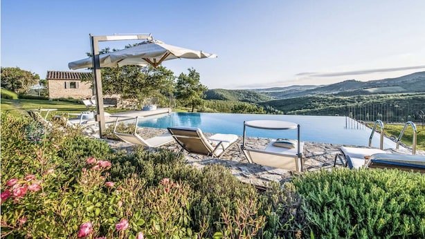 Ein unendlicher Pool mit Liegestühlen und einem Tisch, umgeben von üppiger Vegetation und einem Steinhaus.