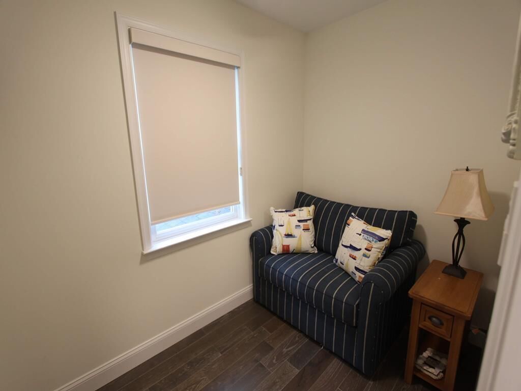 A room with a striped sofa, a small wooden nightstand, a lamp, and a window with blinds.