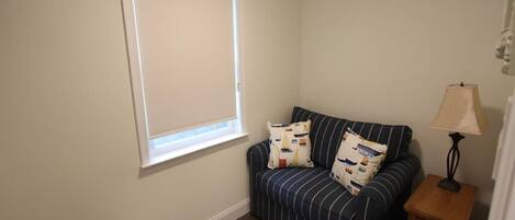 A room with a striped sofa, a small wooden nightstand, a lamp, and a window with blinds.