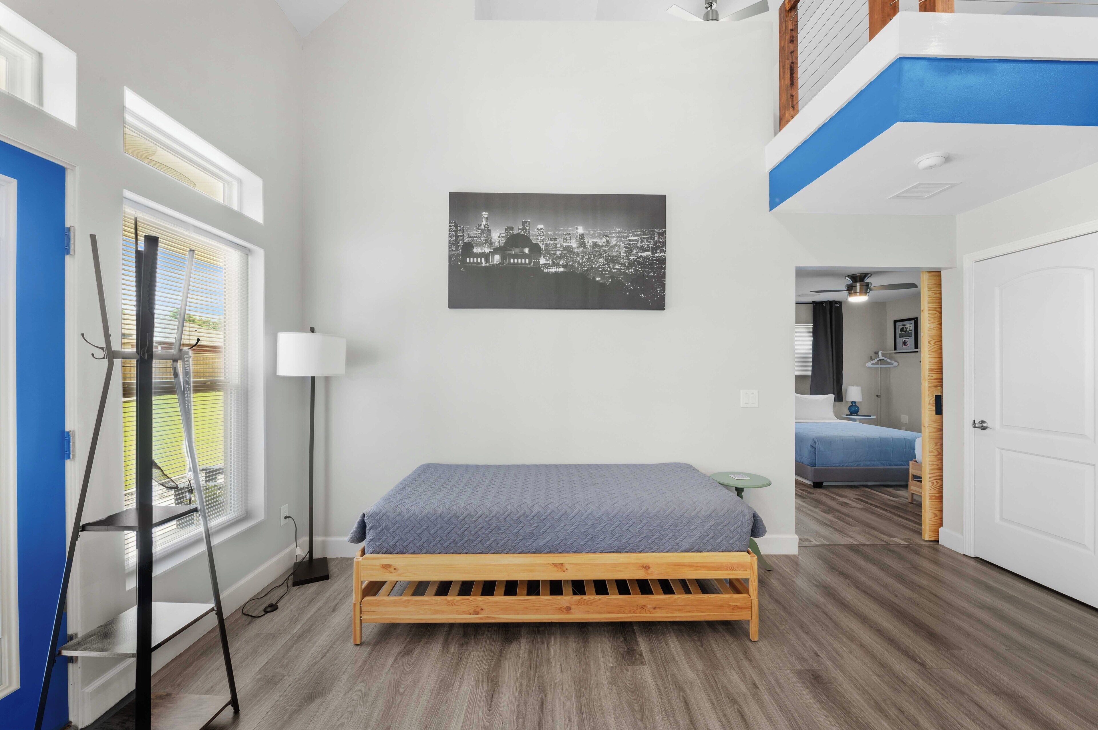 A bedroom with a bed, a cityscape painting, a window with blinds, and a ceiling fan.