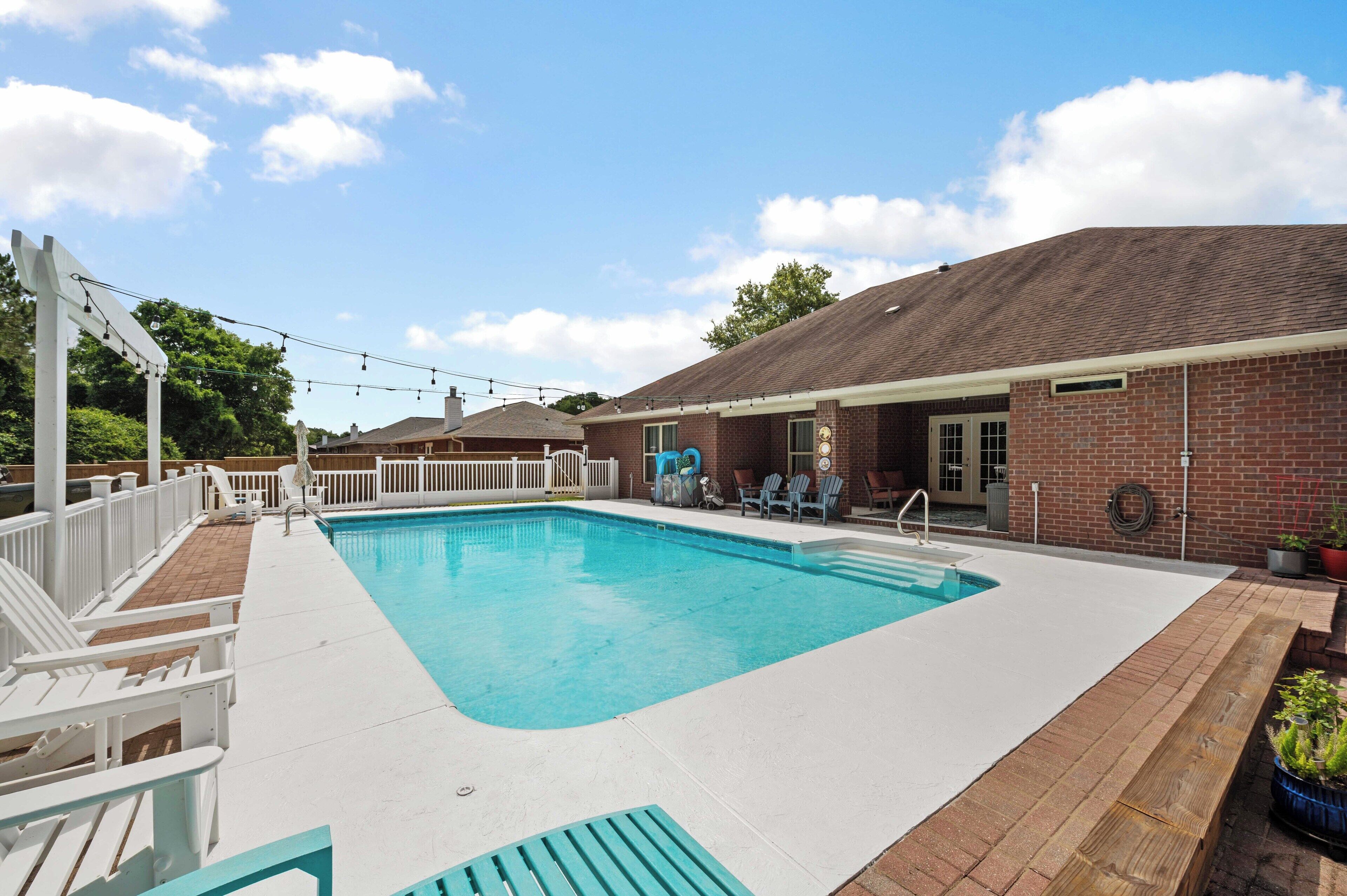 A rectangular swimming pool with a white deck, surrounded by a white fence and several lounge chairs.