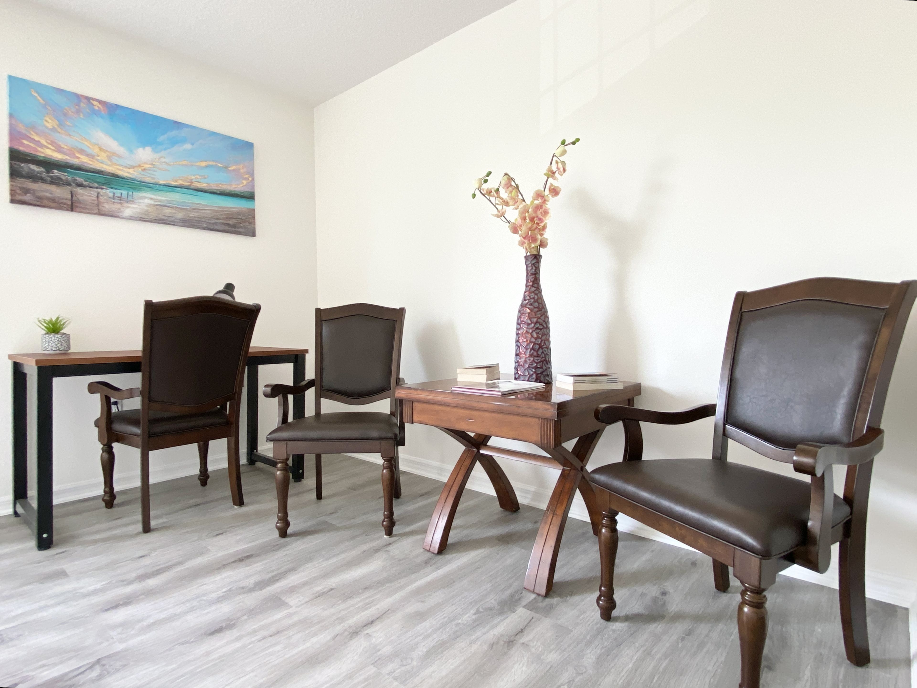 A room with a wooden desk, two chairs, a vase with flowers, and a painting on the wall.