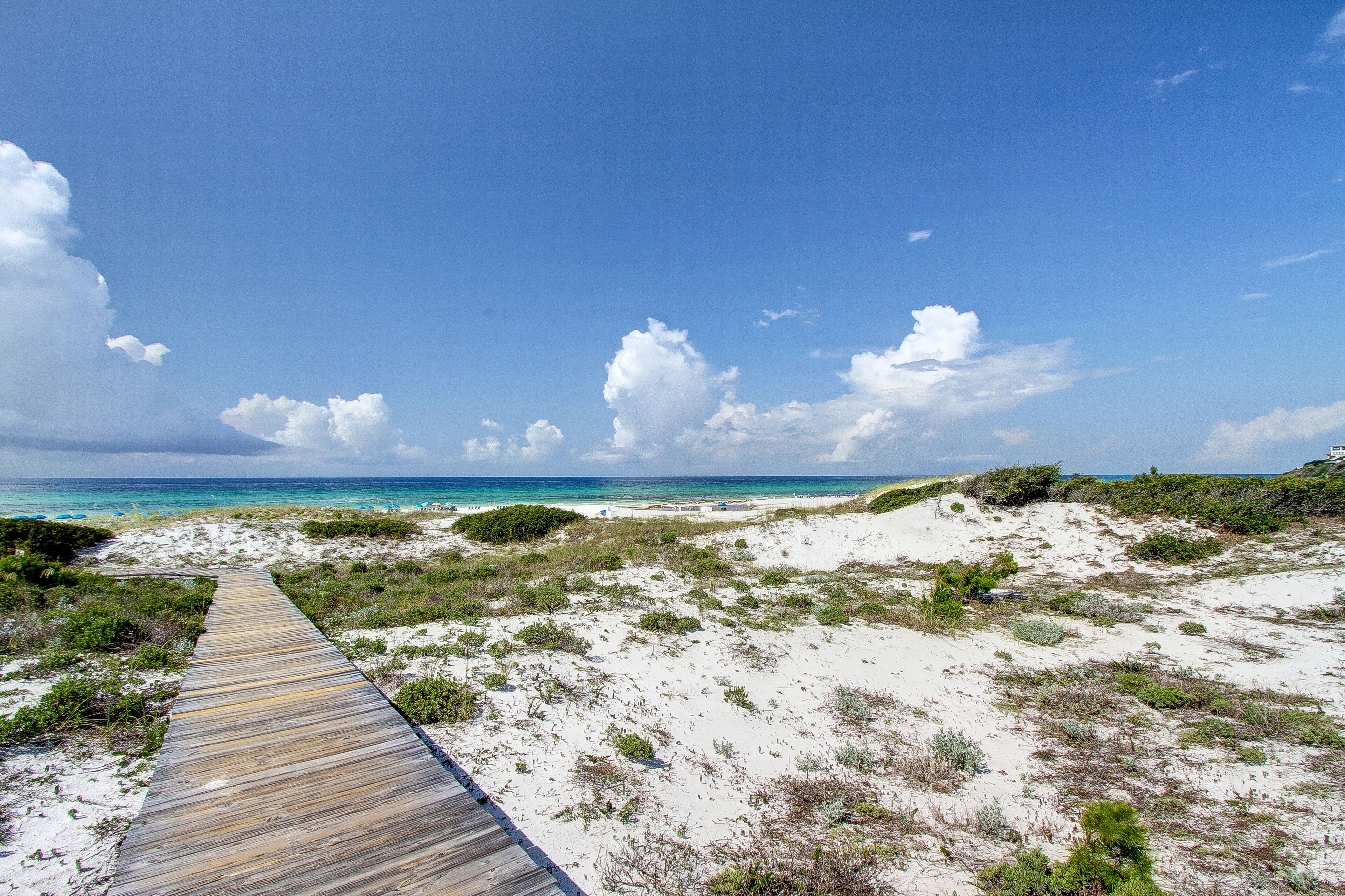A wooden boardwalk leading to a beach with white sand and sparse vegetation, under a clear blue sky.