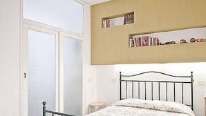 Ein Schlafzimmer mit einem Einzelbett, einem Fenster mit Jalousien, einem Bücherregal mit Büchern und einem kleinen Nachttisch.