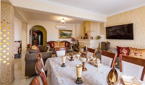 A dining area with a table set for four, surrounded by ornate chairs. A fireplace and a painting are visible in the background.