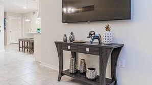A modern living room with a dark wooden console table, a flat-screen TV mounted above it, and a kitchen area in the background. - Spacious End-Unit Condo | Modern Open Floor Plan w/ Elegant Décor & Full Comfort | Minutes to Disney (Kissimmee)