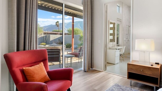 Een moderne hotelkamer met een grote schuifdeur naar een terras, een rode fauteuil, een houten nachtkastje en een zichtbare badkamer door de deur.