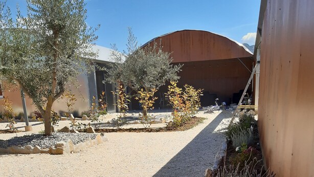 Un chemin de gravier mène à un bâtiment doté d’un toit incurvé, qui est entouré de différentes plantes et d’arbres.