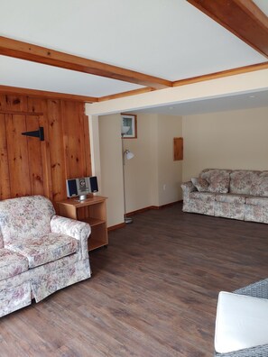 A living room with wooden walls, a wooden ceiling, two floral-patterned sofas, a small wooden table with a lamp, and a television. - Beautiful Lake Winnipesaukee Waterfront! (Alton)