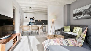 Una sala moderna con sofá, una mesa de centro y un mueble para televisión. Hay un comedor con sillas y una mesa. La habitación tiene un techo con vigas a la vista y un ventanal grande.