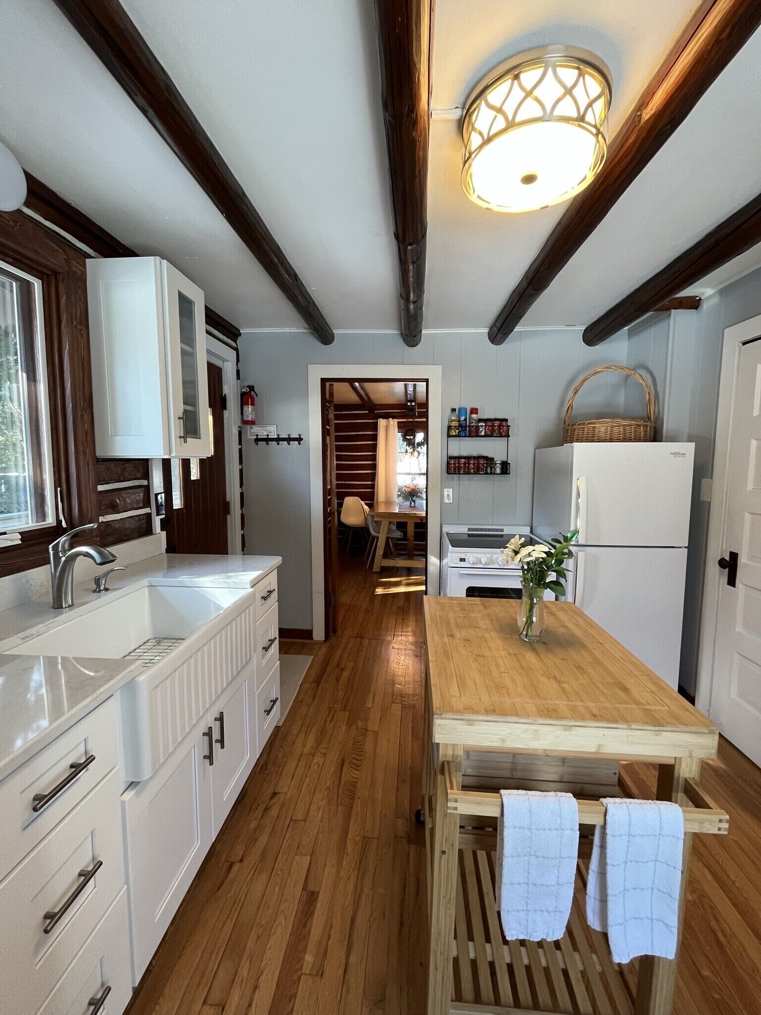 Una cocina con vigas de madera, armarios blancos, una mesa de comedor de madera y una lámpara colgante.