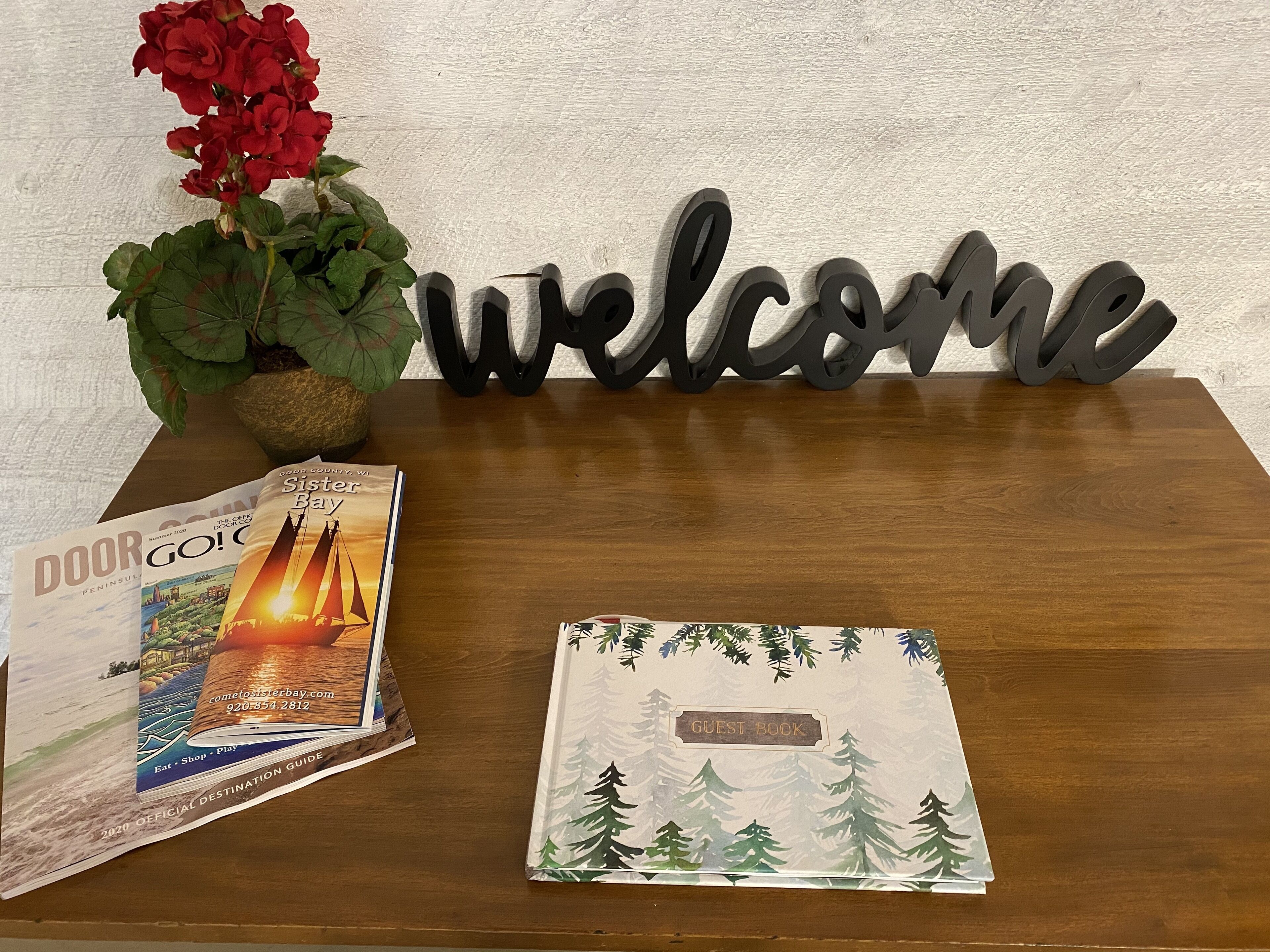 A wooden table with a welcome sign, a guest book, a brochure, and a potted plant.