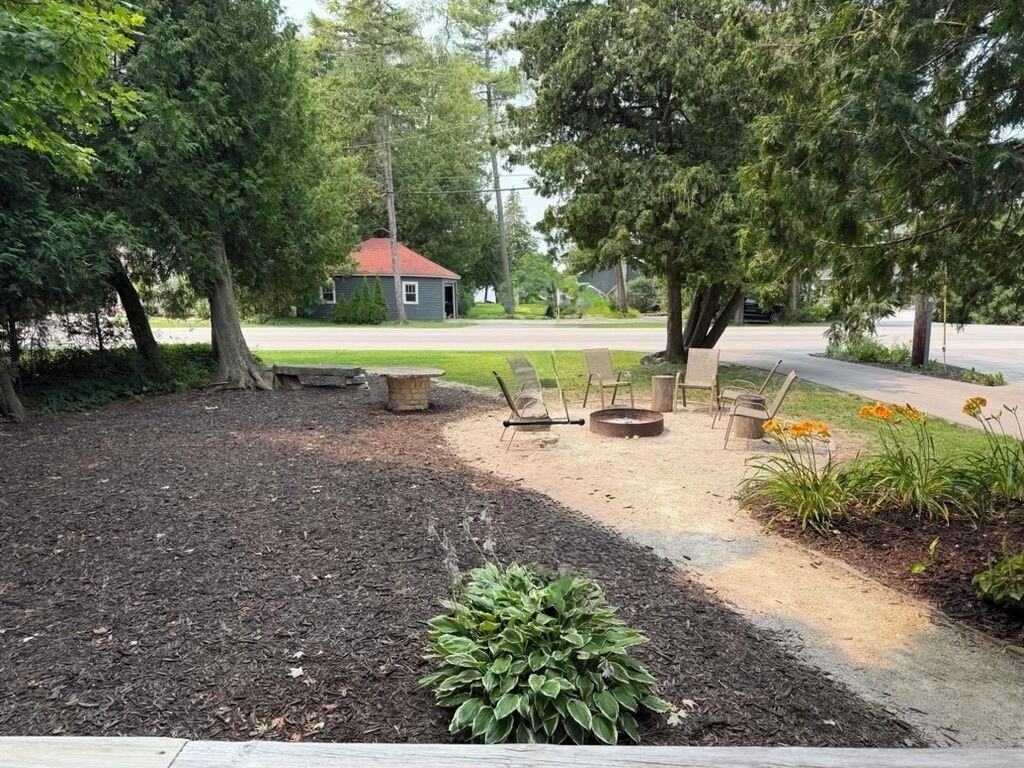 An outdoor seating area with a fire pit, surrounded by trees and a small building in the background.