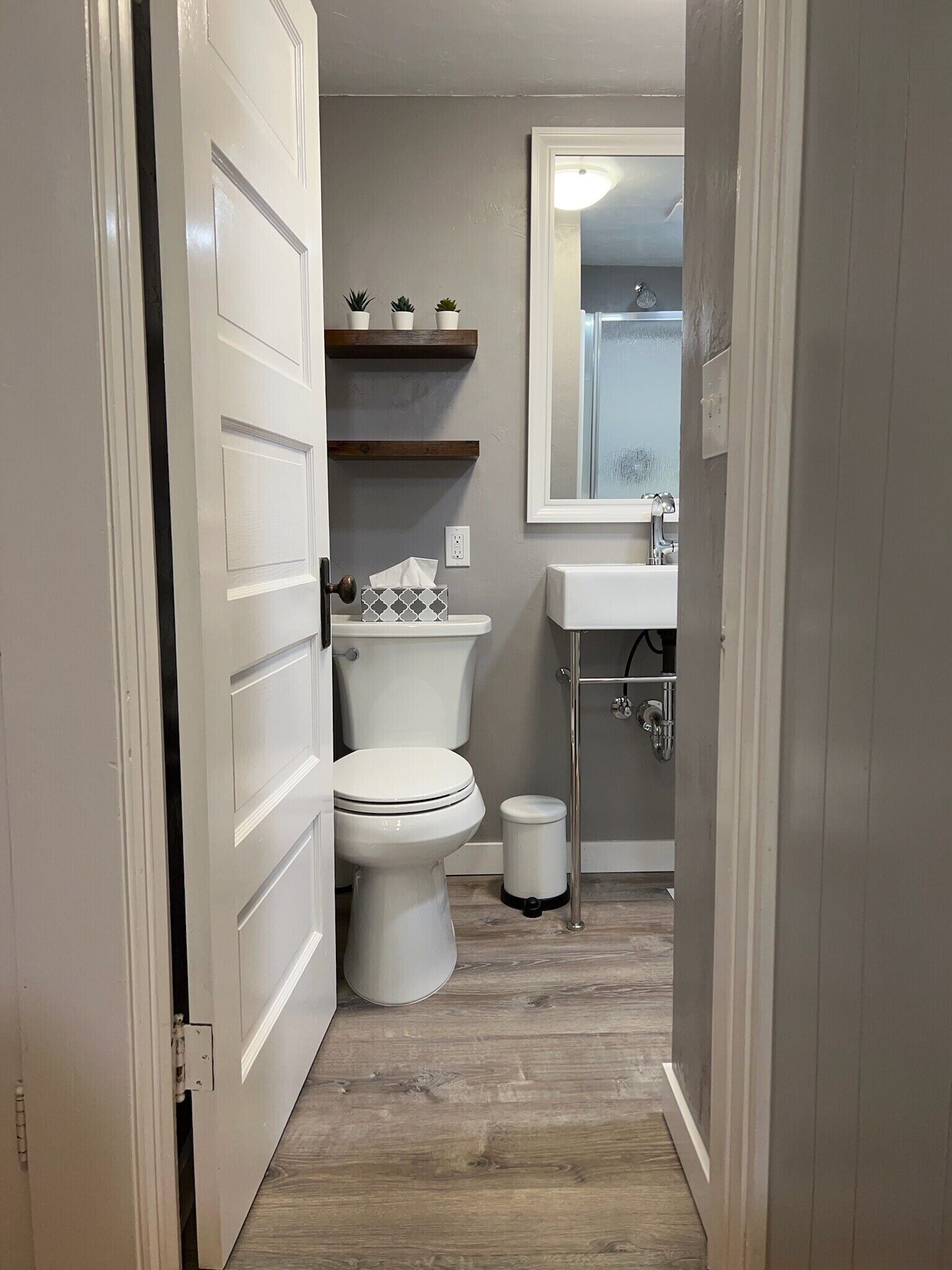 Een compacte badkamer met toilet, wastafel en spiegel. Boven het toilet zijn planken en naast de wastafel een prullenbak.